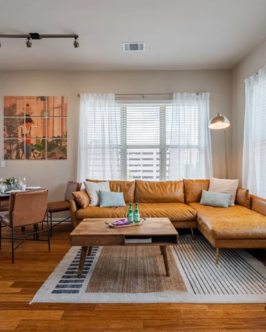 Bright open-plan living room with tan leather sectional sofa, wooden coffee table, dining area, and floor-to-ceiling windows with sheer curtains overlooking