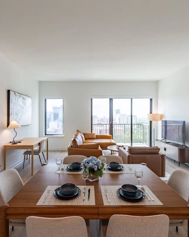 Open-plan apartment with dining table, caramel leather sofa, and floor-to-ceiling windows overlooking city skyline