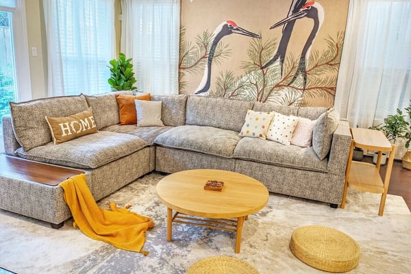 Living room with gray sectional sofa, round wooden coffee table, yellow throw blanket, and wall mural of two cranes with tall grass
