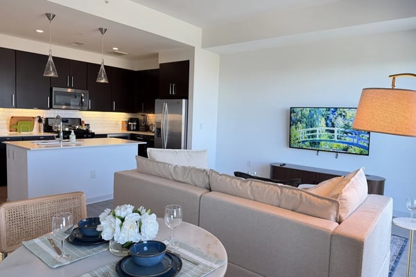 Open-plan living room and kitchen with gray sofa, dining table, dark cabinetry, stainless steel appliances, and wall-mounted TV