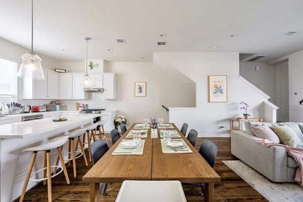 Open-plan kitchen, dining, and living area with white cabinetry, wood dining table set for six, and gray sofa with staircase visible in background.