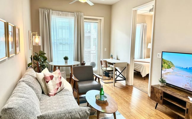 Modern studio apartment with gray sofa, work desk, TV, and floor-to-ceiling windows overlooking city skyline